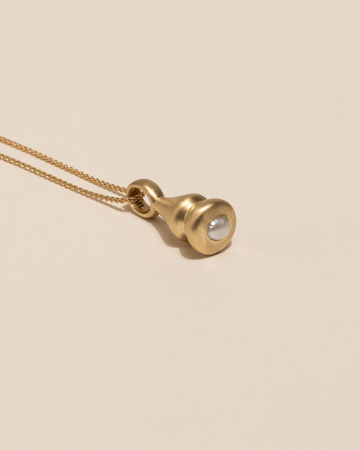 A gold pendant necklace featuring a small, bulbous gold charm with a smooth, round pearl bezel set, resting on a warm beige background.