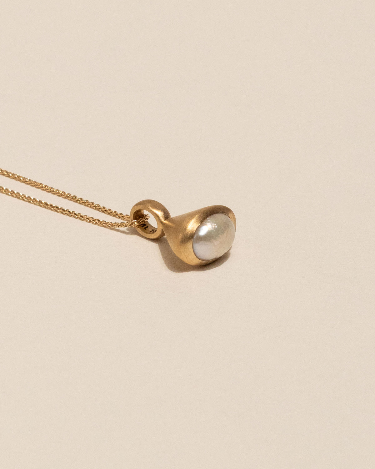 A gold pendant necklace featuring a bell shaped gold charm with a smooth, oval pearl bezel set, resting on a warm beige background.