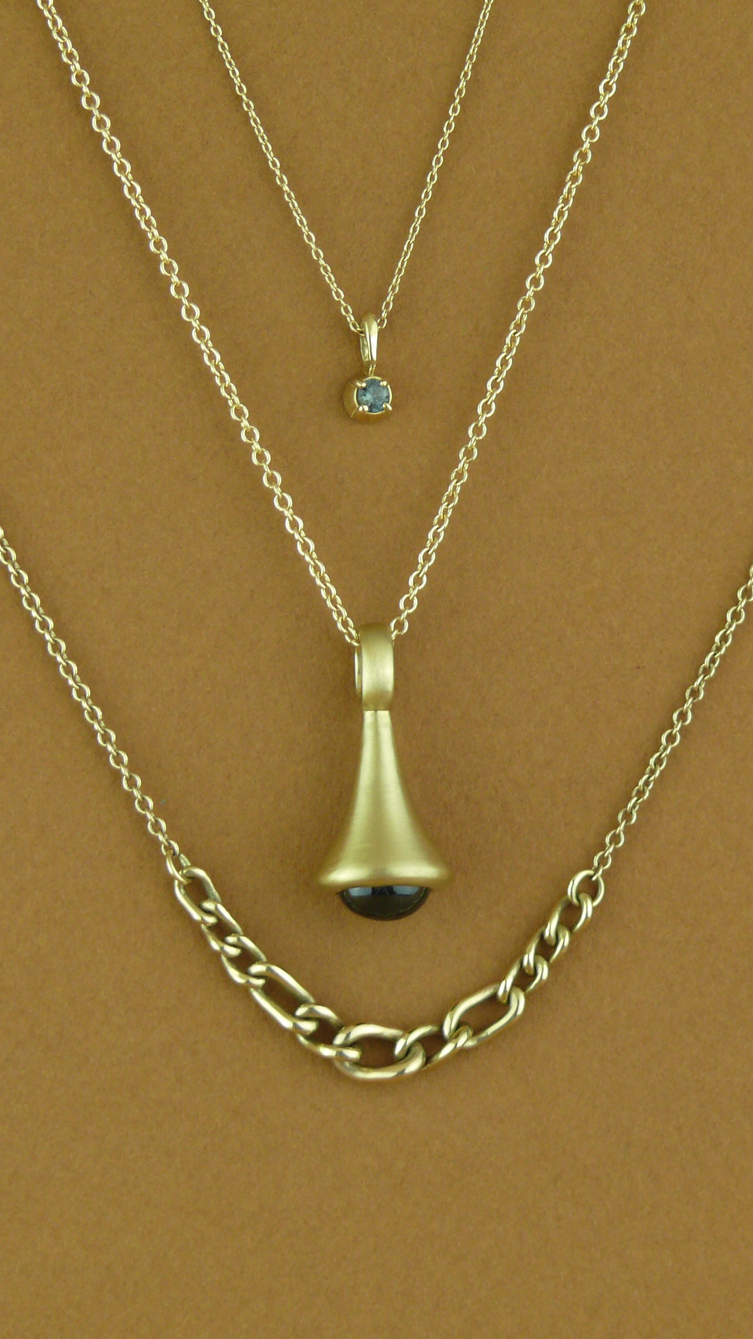 One-of-a-Kind Gold Blue Sapphire Pendulum Charm No. 2, graduated curb chain necklace, and tourmaline blossom charm on brown paper