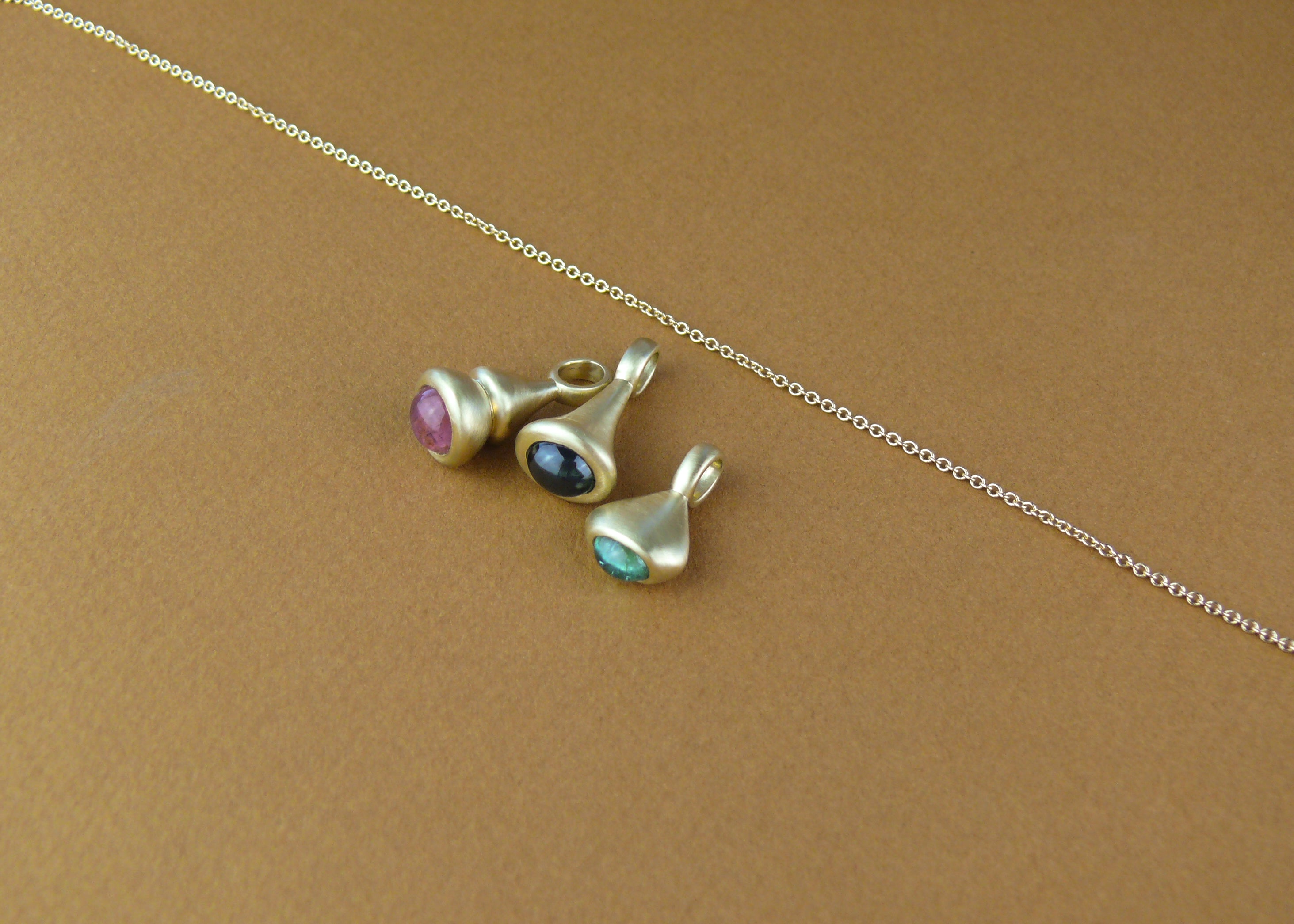 Gold Pendulum Charms with pink tourmaline, blue sapphire, and teal tourmaline on brown paper