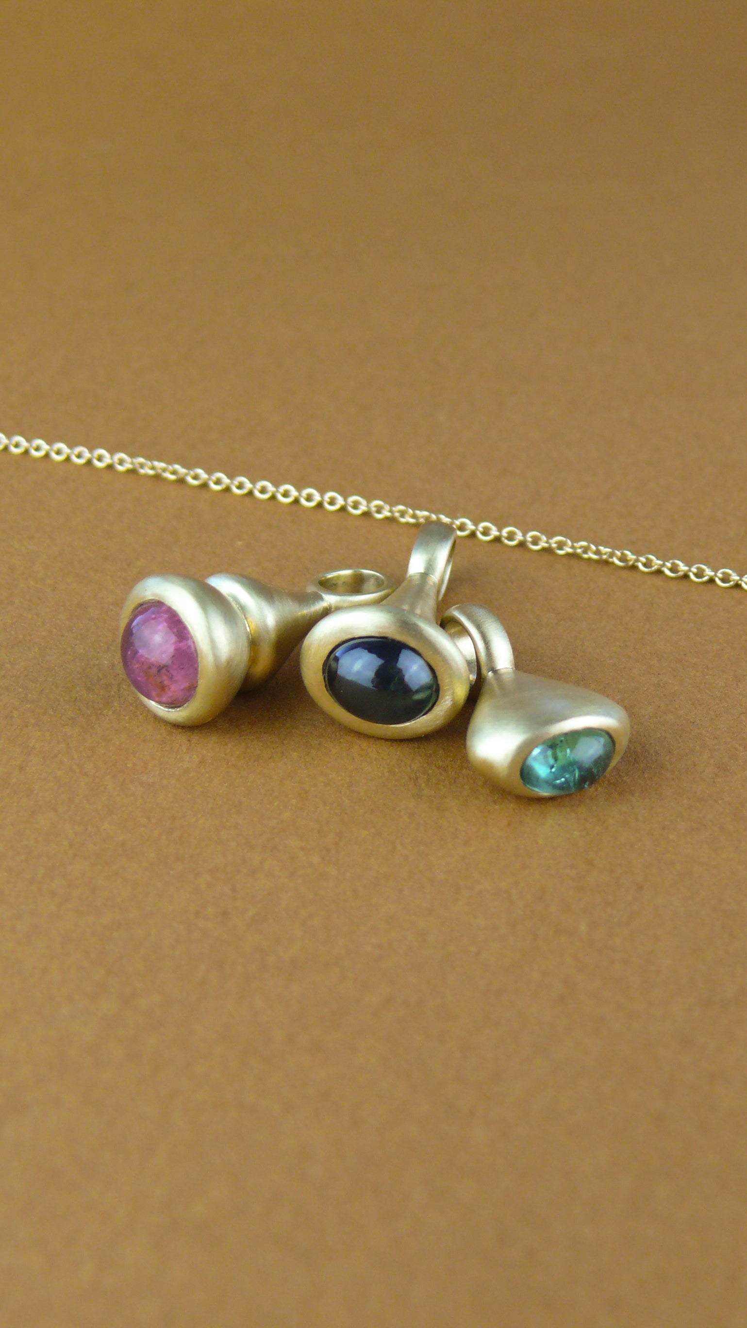 Gold Pendulum Charms with pink tourmaline, blue sapphire, and teal tourmaline on brown paper