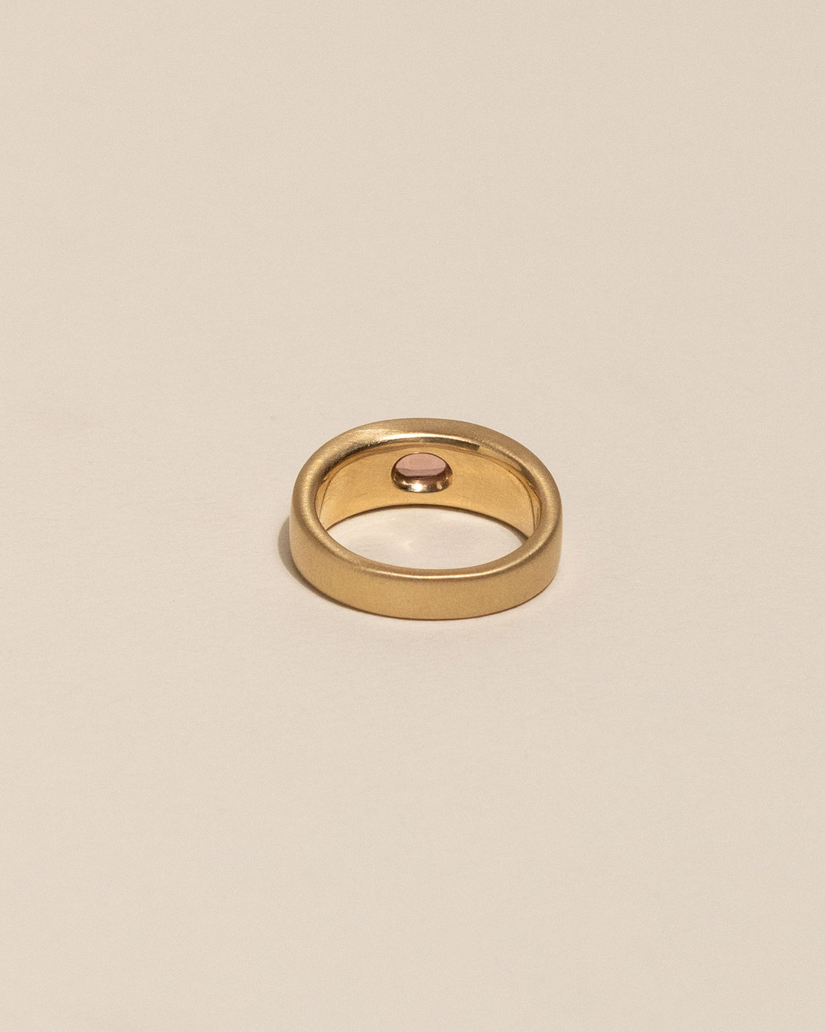 The back of a gold plateau ring with a small oval pink buff top sapphire flush set into a flat gold band, centered on a plain beige background.
