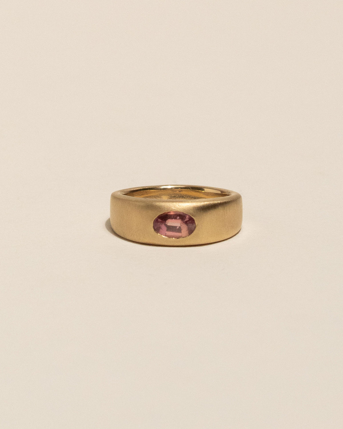 A gold plateau ring with a small oval pink buff top sapphire flush set into a flat gold band, centered on a plain beige background.
