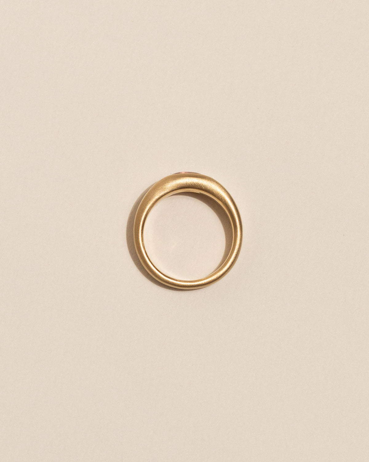 A gold plateau ring with a small oval pink buff top sapphire flush set into a flat gold band, centered on a plain beige background.
