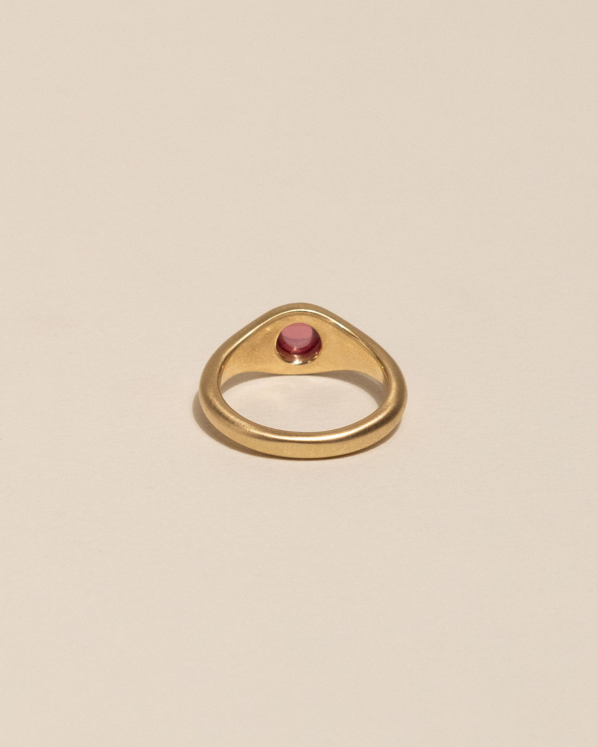 Back of a gold signet-style ring with a smooth, rounded pink gemstone in the center, displayed on a clean, solid beige background.