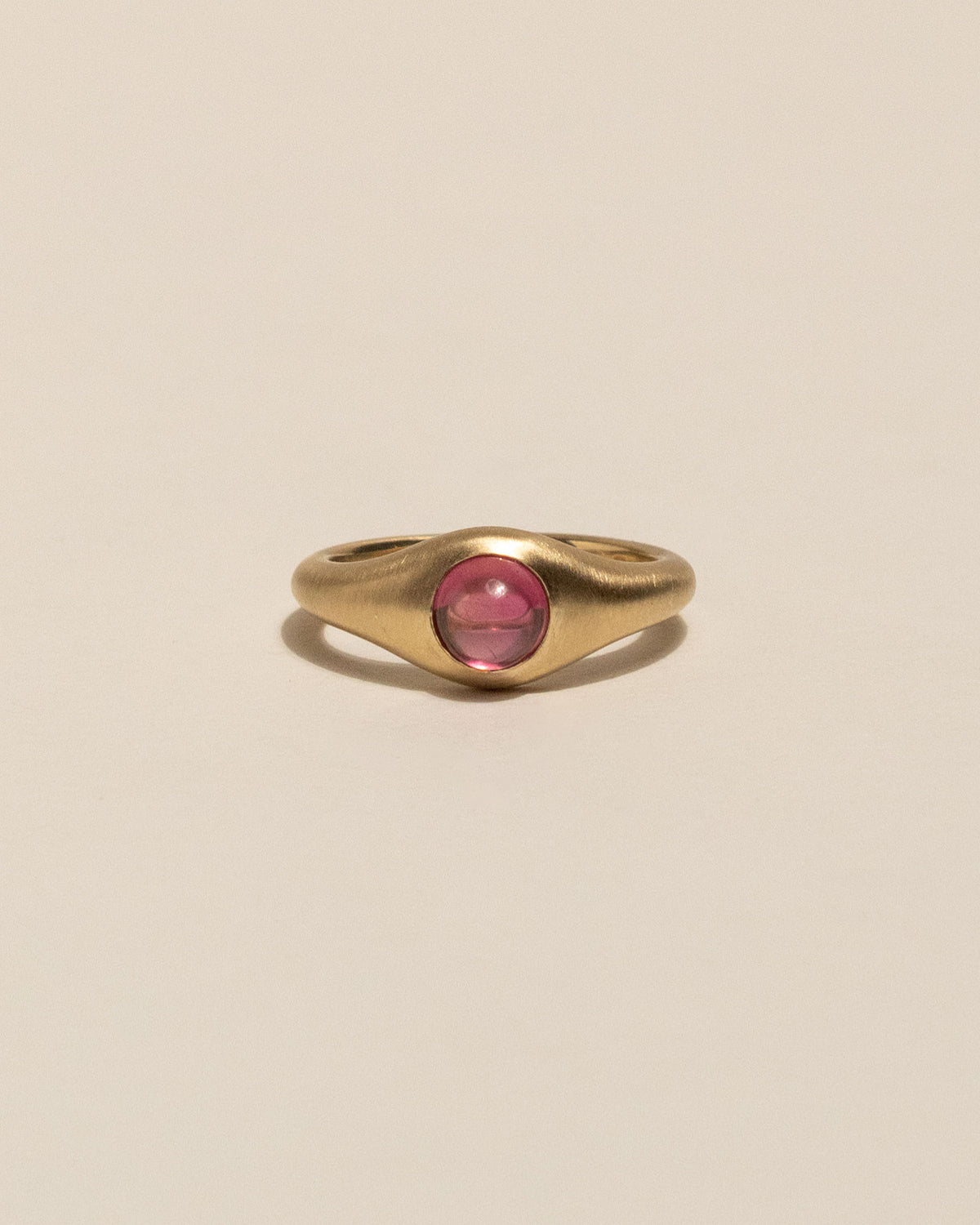 A gold signet-style ring with a smooth, rounded pink gemstone in the center, displayed on a clean, solid beige background.