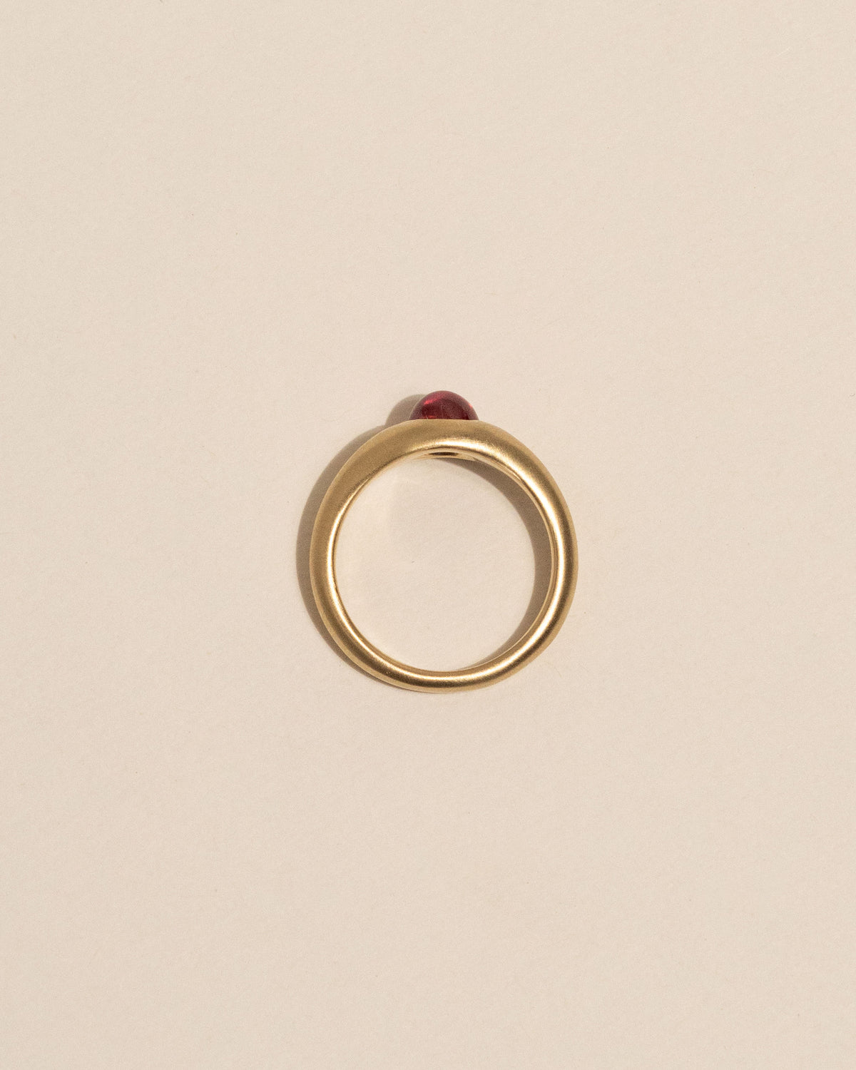 Top view of a gold signet-style ring with a smooth, rounded pink gemstone in the center, displayed on a clean, solid beige background.
