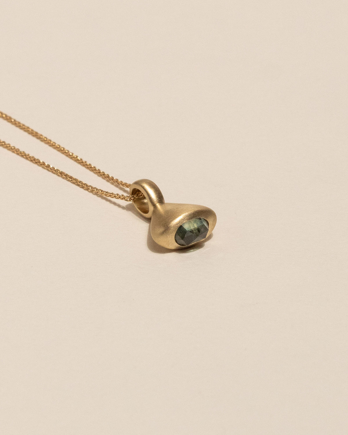A gold pendant necklace featuring a small, bell shaped gold charm with a rose cut bi-colored Montana sapphire bezel set, resting on a warm beige background.