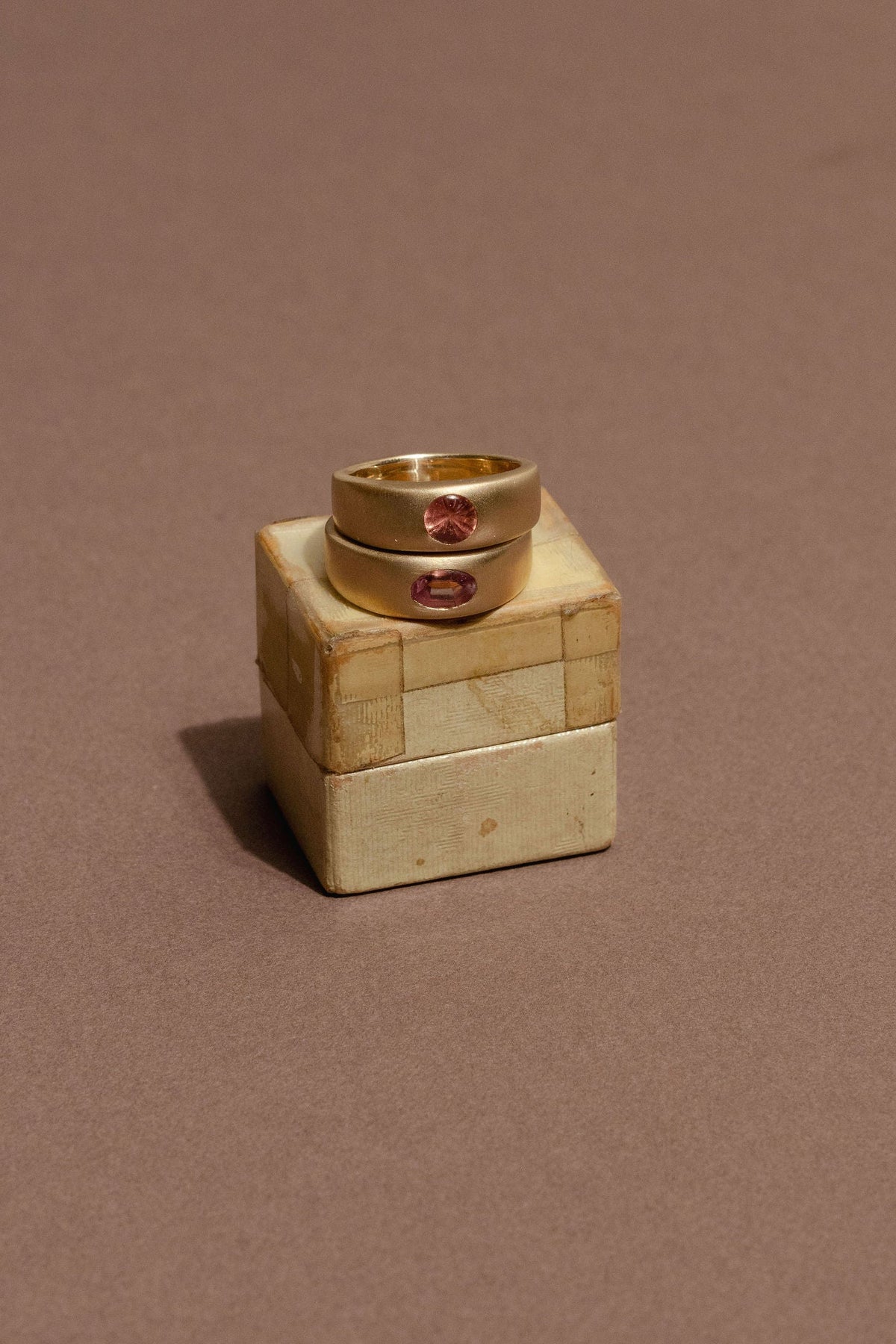 Two gold rings with small pink gemstone accents displayed on a small beige block against a solid brown background.