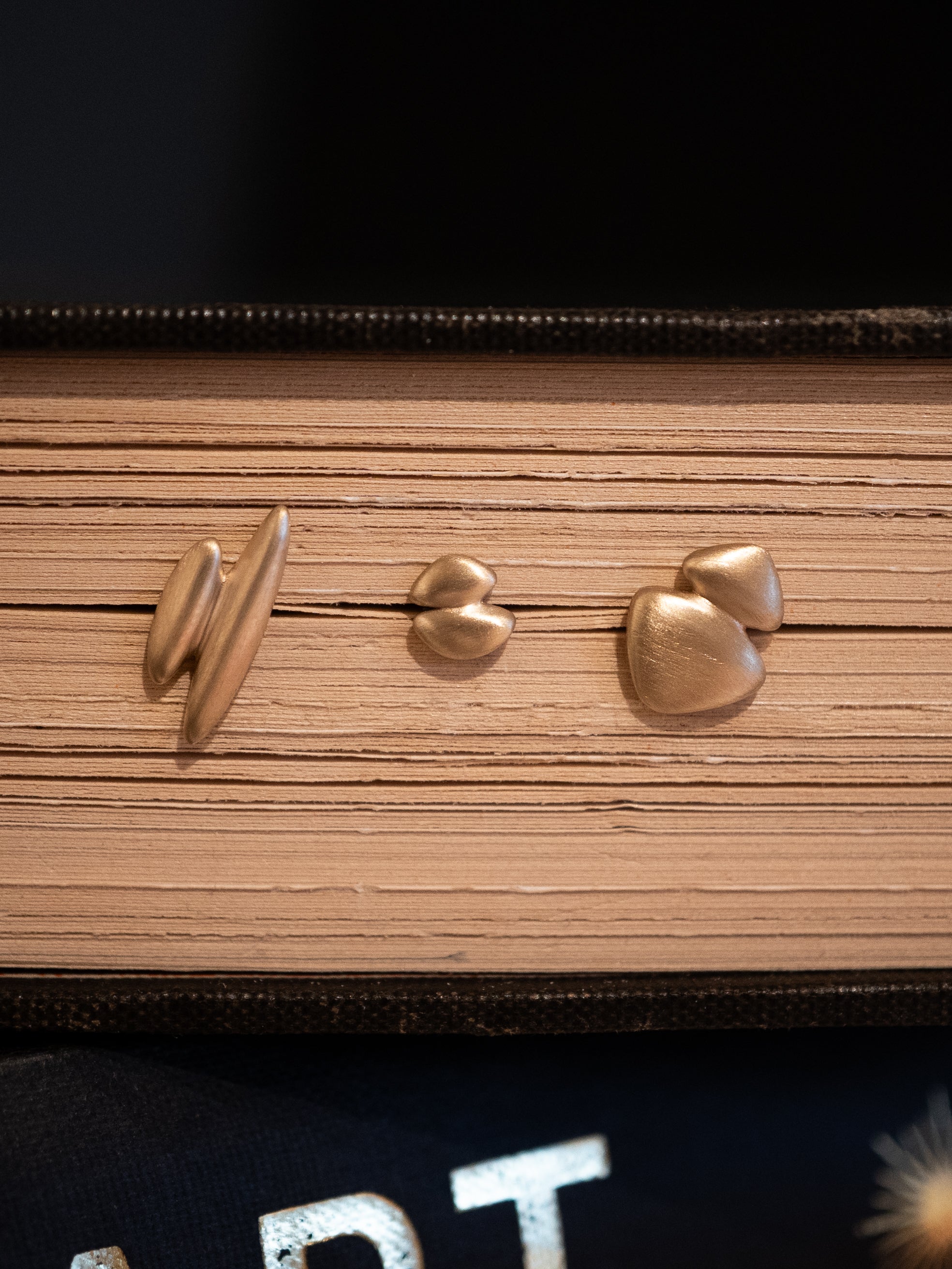 Gold stud earrings with matte texture and organic forms on a book surface.