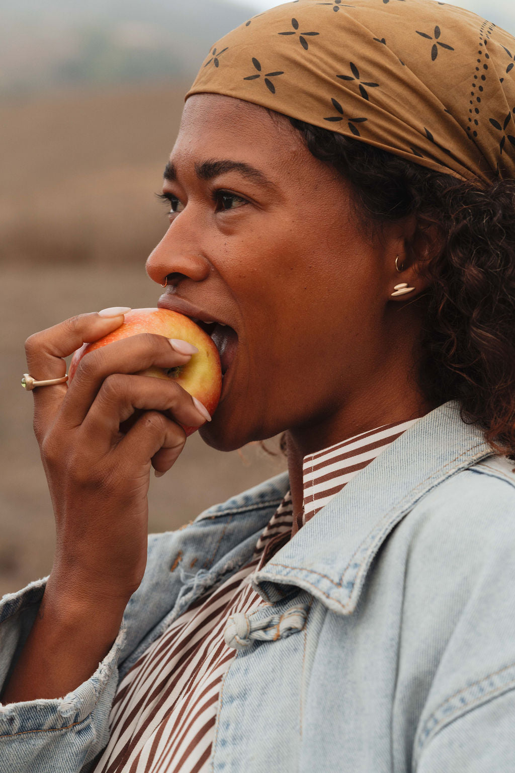 Model with bandana on her head eating and apple and wearing gold earrings and rings from the Made Line Jewelry Trove Collection. 
