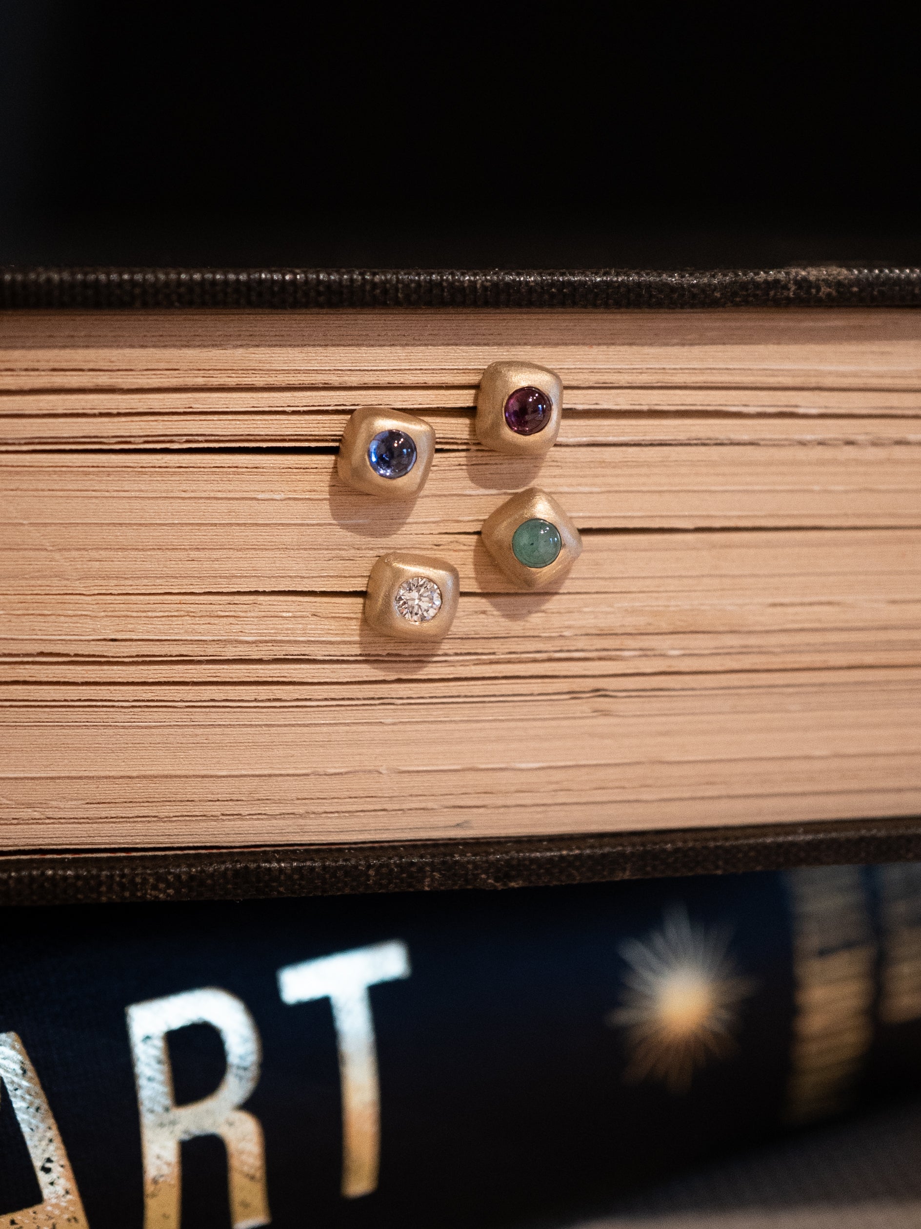 Four gold Trove Stud earrings in the shape of soft squares, with a diamond, sapphire, garnet, and emerald, placed on a book background. 