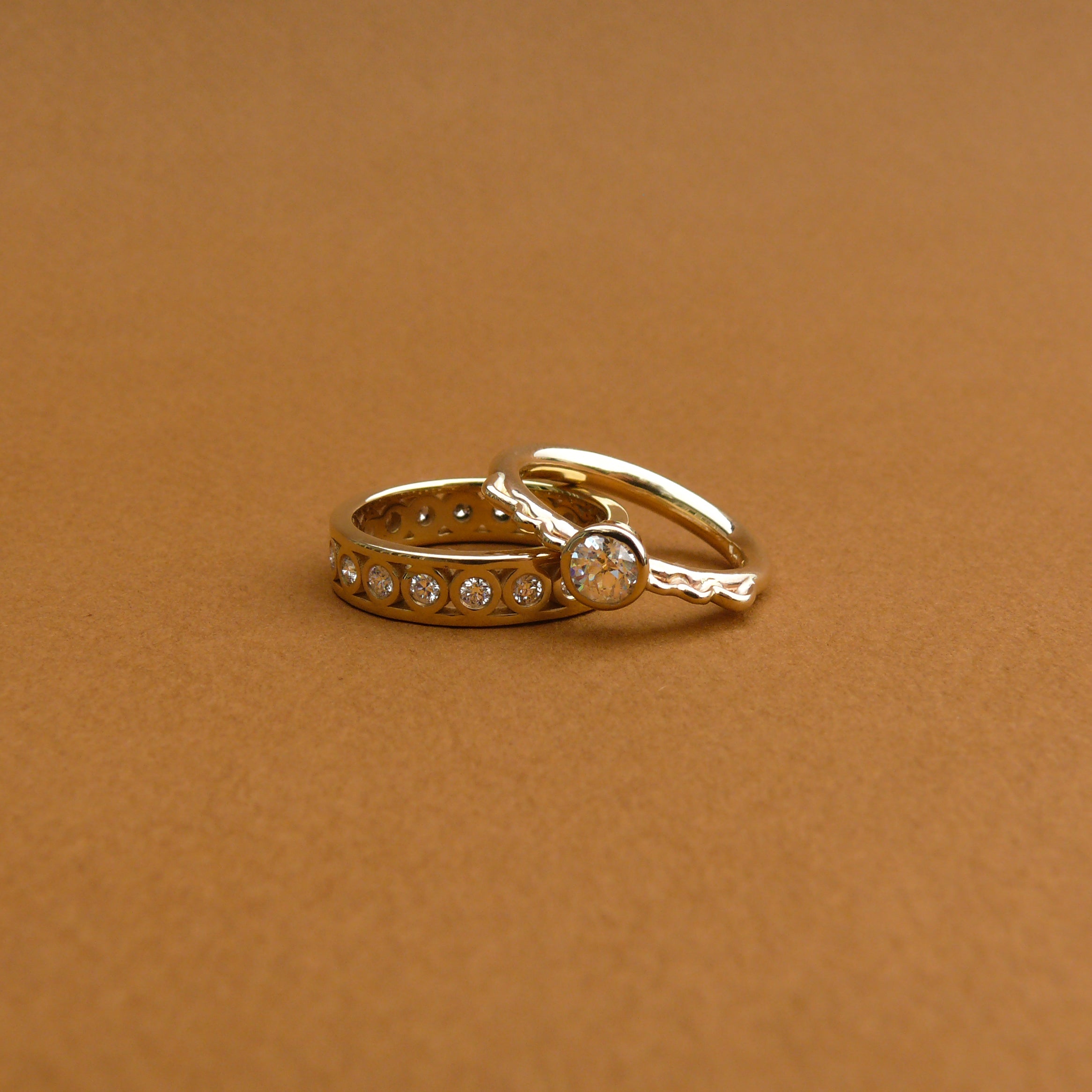 Two rings, all white diamond bezel set on gold rings that have the profile of a vessel. One ring is a diamond eternity band.