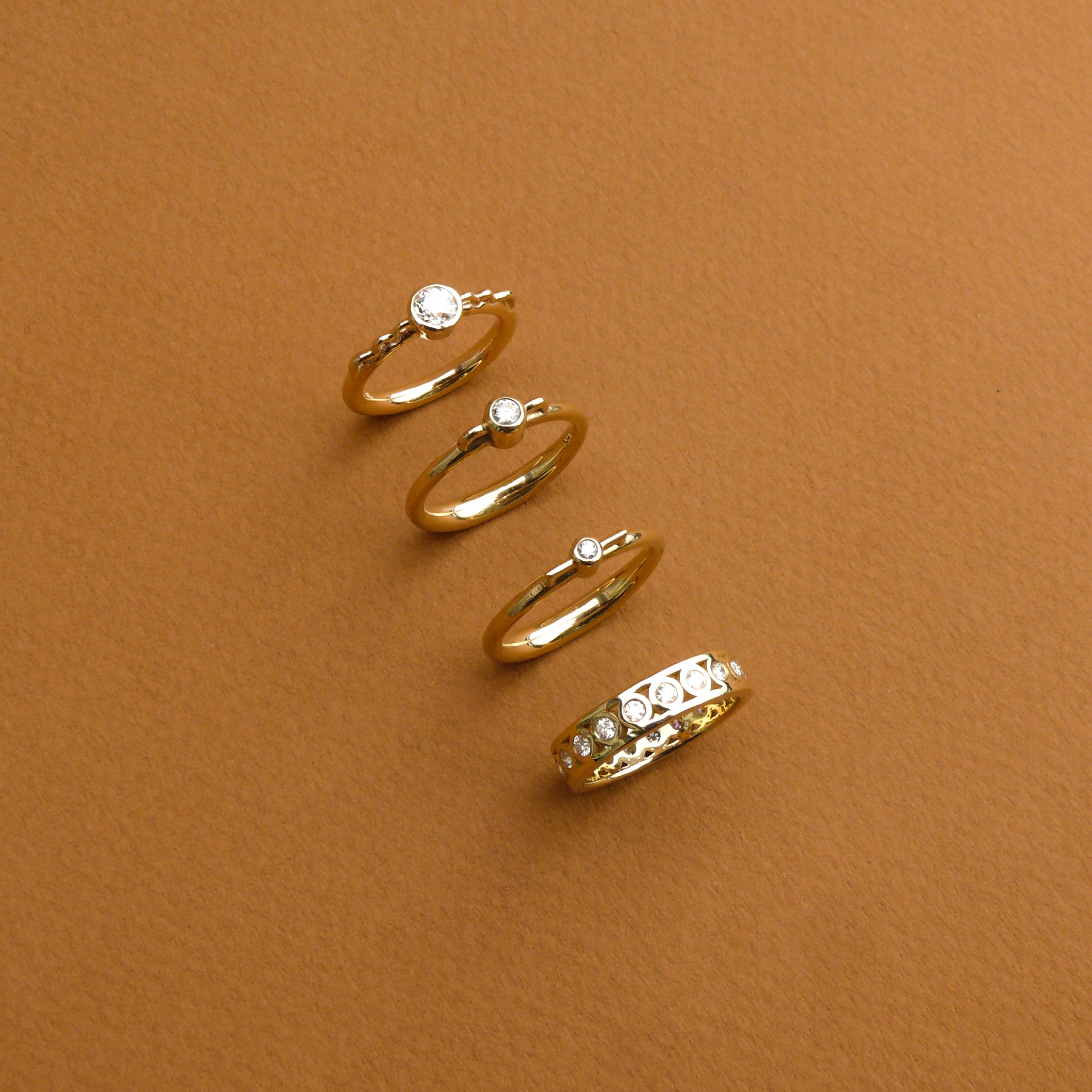 Four rings, all white diamond bezel set on gold rings that have the profile of a vessel. One ring is a diamond eternity band.